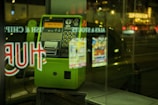 A green public payphone is situated behind a glass window displaying reflections of lit-up signs from a street at night. The payphone has a numeric keypad, coin slot, and labels in Japanese. The background contains blurred city lights with vehicles passing by.