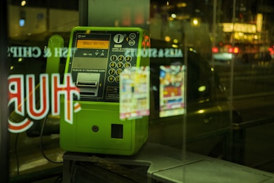 A green public payphone is situated behind a glass window displaying reflections of lit-up signs from a street at night. The payphone has a numeric keypad, coin slot, and labels in Japanese. The background contains blurred city lights with vehicles passing by.