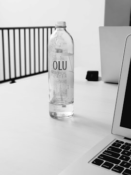 A clear bottle labeled 'OLU Tropical Water' is placed on a white tabletop. In the background, there are parts of a laptop, a power adapter, and a railing. The setting appears minimalistic with a focus on the water bottle.