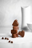 A stack of smooth, heart-shaped chocolates sits elegantly on a textured white surface. The background is softly blurred, with subtle tones that enhance the focus on the rich, brown chocolates. Additional chocolate pieces are scattered nearby, adding to the composition.