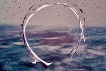 An abstract image featuring a swirling circular brushstroke in white against a textured background. The backdrop appears to be an artistic representation of water, with splashes of purple and pink dots scattered throughout.