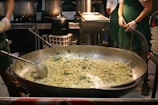 A large pan filled with tortellini pasta being prepared in a busy kitchen setting. Several chefs wearing green aprons and gloves are working around the cooking station, with various pots and containers visible in the background.