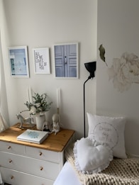 A cozy bedroom corner featuring a white dresser topped with decorative items including a vase of flowers, a rocking horse, and plush toys. Above the dresser hang three framed pictures, one with text. The wall also has a large floral decal. A lamp stands to the side, and a bed with pillows, including a heart-shaped one, has a knitted throw.