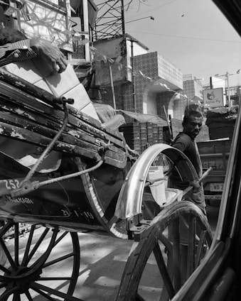 A black and white image depicting a man next to a rickshaw on a busy street. The rickshaw carries a number of stacked boxes. In the background, various buildings and a sign reading 'Saha Ply' are visible. The man's expression seems focused as he stands near the vehicle.