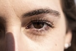 A close-up view of a person's eye with detailed focus on the eyelashes and eyebrow. The lighting highlights the texture of the skin around the eye, casting a subtle shadow.