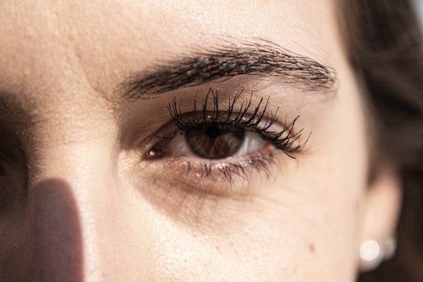 A close-up view of a person's eye with detailed focus on the eyelashes and eyebrow. The lighting highlights the texture of the skin around the eye, casting a subtle shadow.