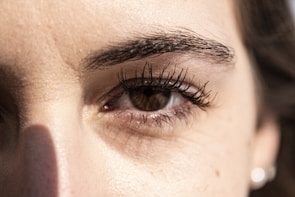 A close-up view of a person's eye with detailed focus on the eyelashes and eyebrow. The lighting highlights the texture of the skin around the eye, casting a subtle shadow.