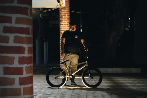 A person stands next to a BMX bicycle in a dimly lit area with brick columns. The person is illuminated by a focused light source, creating contrast with the darker surroundings.