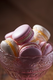 A collection of pastel-colored macarons arranged in an ornate glass dish with intricate floral patterns. The macarons are primarily pink and white, with a smooth texture and creamy fillings.