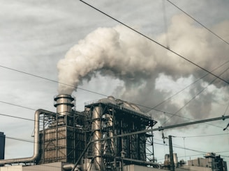 A large industrial facility with metal structures and chimneys emitting thick clouds of smoke into a cloudy sky. Electrical wires intersect the scene, adding to the industrial atmosphere.