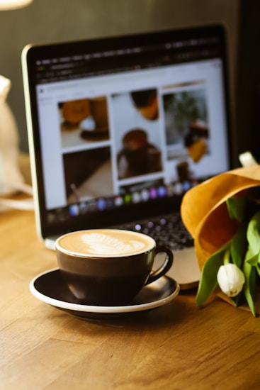A cup of coffee with latte art sits on a saucer in the foreground, placed on a wooden table. In the background, a laptop displays a blurred web page with several images. A bouquet of white tulips is partially visible beside the cup, adding a touch of freshness to the scene.
