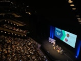 A large audience is seated in a dimly lit auditorium, watching a panel discussion on stage. Four people are seated in chairs around small tables, with a large screen displaying information about the panelists and the topic 'Creating and Managing Scalable Class Products'. The hashtag '#PRODUCTCON' is prominently displayed on the stage.