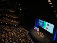 A large audience is seated in a dimly lit auditorium, watching a panel discussion on stage. Four people are seated in chairs around small tables, with a large screen displaying information about the panelists and the topic 'Creating and Managing Scalable Class Products'. The hashtag '#PRODUCTCON' is prominently displayed on the stage.