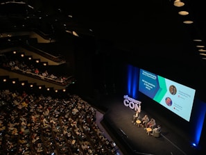 A large audience is seated in a dimly lit auditorium, watching a panel discussion on stage. Four people are seated in chairs around small tables, with a large screen displaying information about the panelists and the topic 'Creating and Managing Scalable Class Products'. The hashtag '#PRODUCTCON' is prominently displayed on the stage.