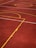 A sports court with a reddish-brown surface and bright yellow and white lines. The lines form geometric shapes, including rectangles and circles, suggesting markings for games like basketball or netball.