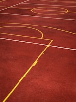 A sports court with a reddish-brown surface and bright yellow and white lines. The lines form geometric shapes, including rectangles and circles, suggesting markings for games like basketball or netball.