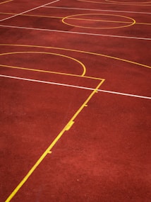 A sports court with a reddish-brown surface and bright yellow and white lines. The lines form geometric shapes, including rectangles and circles, suggesting markings for games like basketball or netball.