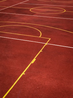 A sports court with a reddish-brown surface and bright yellow and white lines. The lines form geometric shapes, including rectangles and circles, suggesting markings for games like basketball or netball.