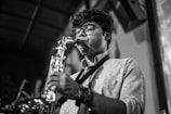 A person wearing glasses plays a saxophone with focused concentration. The setting appears to be indoors, possibly in a performance venue, as indicated by the blurred background and stage-like environment.