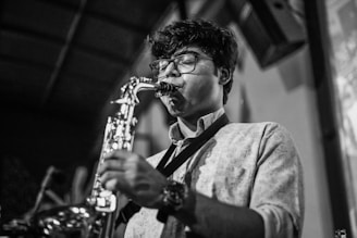 A person wearing glasses plays a saxophone with focused concentration. The setting appears to be indoors, possibly in a performance venue, as indicated by the blurred background and stage-like environment.