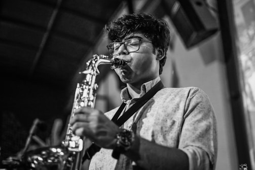 A person wearing glasses plays a saxophone with focused concentration. The setting appears to be indoors, possibly in a performance venue, as indicated by the blurred background and stage-like environment.