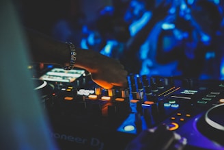 A DJ is adjusting the controls on a mixing console, surrounded by vibrant lights that create a dynamic atmosphere. The DJ's hand is in focus, with details of various buttons and knobs visible on the console. The background is blurred, with blue lights and silhouettes of people suggesting a lively crowd.