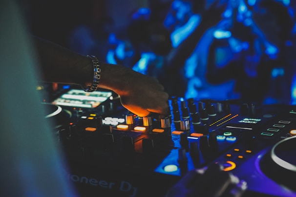 A DJ is adjusting the controls on a mixing console, surrounded by vibrant lights that create a dynamic atmosphere. The DJ's hand is in focus, with details of various buttons and knobs visible on the console. The background is blurred, with blue lights and silhouettes of people suggesting a lively crowd.