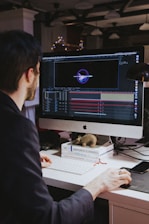A person with dark hair is working at a desk with a large monitor displaying a design or animation software interface. The desk is cluttered with books and a small toy-like object. The background is dimly lit with hanging overhead lights and blurred decorative lights.