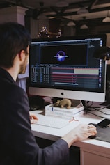A person with dark hair is working at a desk with a large monitor displaying a design or animation software interface. The desk is cluttered with books and a small toy-like object. The background is dimly lit with hanging overhead lights and blurred decorative lights.