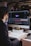 A person with dark hair is working at a desk with a large monitor displaying a design or animation software interface. The desk is cluttered with books and a small toy-like object. The background is dimly lit with hanging overhead lights and blurred decorative lights.
