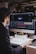 A person with dark hair is working at a desk with a large monitor displaying a design or animation software interface. The desk is cluttered with books and a small toy-like object. The background is dimly lit with hanging overhead lights and blurred decorative lights.