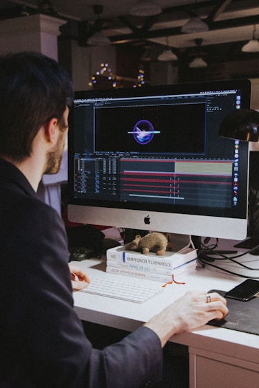A person with dark hair is working at a desk with a large monitor displaying a design or animation software interface. The desk is cluttered with books and a small toy-like object. The background is dimly lit with hanging overhead lights and blurred decorative lights.