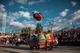 A colorful group of dancers dressed in traditional attire performs an acrobatic move in a public square. The vibrant costumes feature layers of fabric in bright shades like red, green, and yellow. Onlookers gather around, sitting on steps and standing to watch the lively display. The scene is set against a backdrop of a clear, blue sky and scattered clouds, with lampposts and trees framing the area.