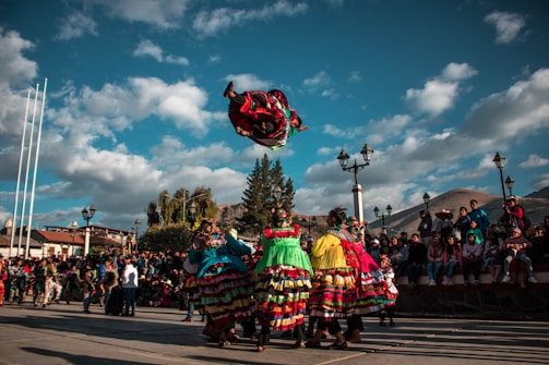 A colorful group of dancers dressed in traditional attire performs an acrobatic move in a public square. The vibrant costumes feature layers of fabric in bright shades like red, green, and yellow. Onlookers gather around, sitting on steps and standing to watch the lively display. The scene is set against a backdrop of a clear, blue sky and scattered clouds, with lampposts and trees framing the area.