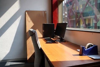 A spacious office desk with two monitors and a cushioned chair. The desk is beside a large window, letting in natural light, and a tape dispenser is visible on the right. A soft glow is cast across the wooden surface, highlighting an organized and minimalist workspace in an urban setting.