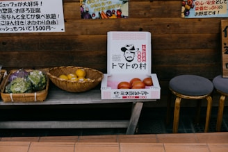 A rustic wooden stall displaying a selection of vegetables and fruits. There are baskets containing leafy greens and what appears to be lemons. A cardboard box with illustrations and text is filled with large tomatoes. The setup is complemented by stools and Japanese signage, contributing to a market-like ambiance.