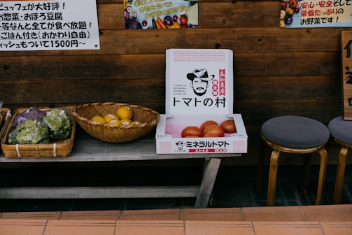 A rustic wooden stall displaying a selection of vegetables and fruits. There are baskets containing leafy greens and what appears to be lemons. A cardboard box with illustrations and text is filled with large tomatoes. The setup is complemented by stools and Japanese signage, contributing to a market-like ambiance.