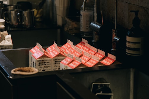 Several milk cartons with open tops are placed in an industrial-style metal sink, surrounded by various kitchen items including bottles and containers. The cartons have red and white labels with text in a foreign language.