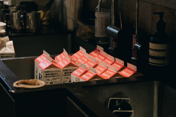 Several milk cartons with open tops are placed in an industrial-style metal sink, surrounded by various kitchen items including bottles and containers. The cartons have red and white labels with text in a foreign language.