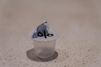 A small, transparent plastic container with a peel-off lid partially opened, placed on a speckled countertop. The lid has some black text printed on it.