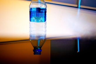 An upside-down plastic water bottle with a blue label is suspended in the foreground. The background consists of a reflective surface with a gradient of warm and cool tones, creating an abstract appearance.