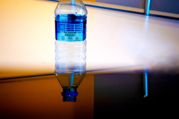 An upside-down plastic water bottle with a blue label is suspended in the foreground. The background consists of a reflective surface with a gradient of warm and cool tones, creating an abstract appearance.