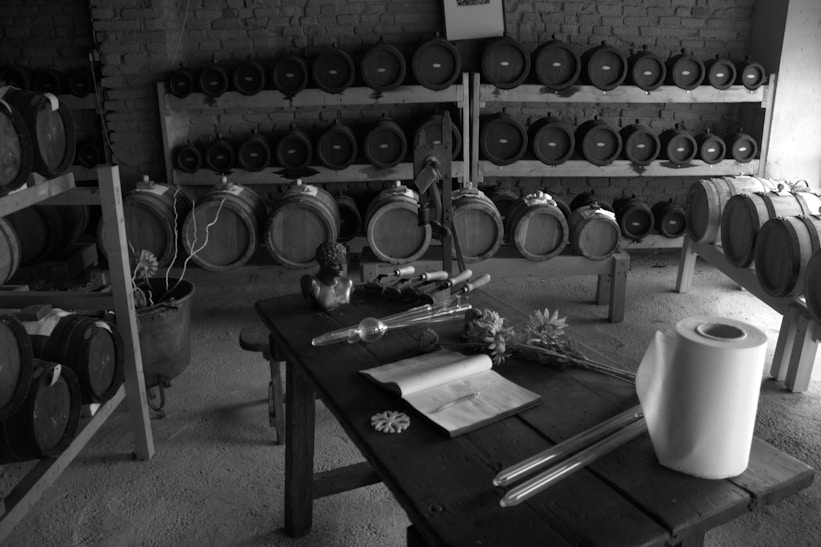 A rustic cellar featuring wooden shelves lined with barrels. A wooden table in the foreground is adorned with a decorative bust, what seems to be writing implements, and scattered flowers. The floor is made of concrete and the walls are brick.