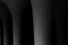A series of curved, dark gray architectural structures creating a sense of depth and shadow. The lighting is minimal, emphasizing the smooth surface texture and creating a dramatic contrast between light and dark areas.