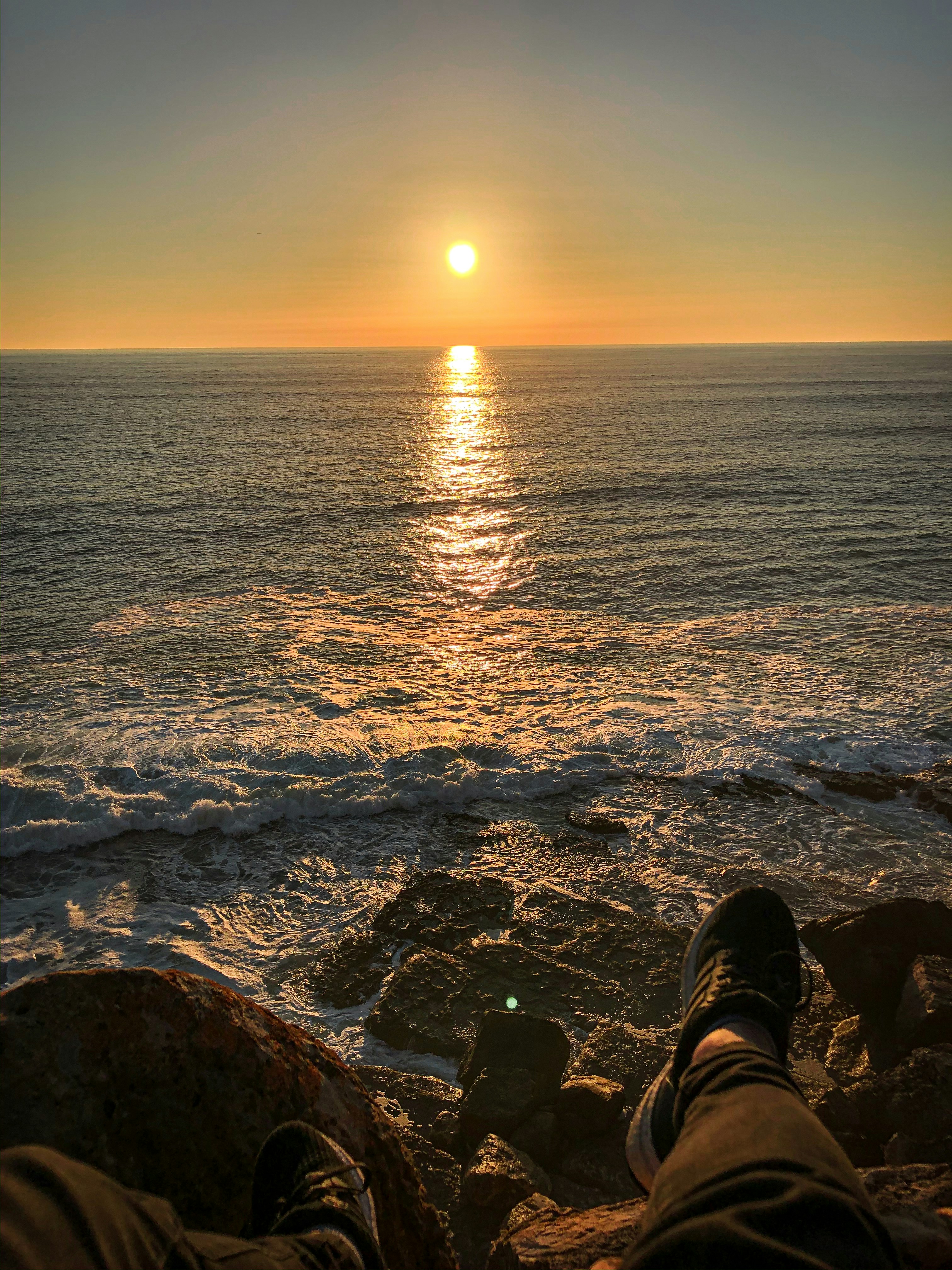 person in black and white sneakers sitting on rock near sea during sunset