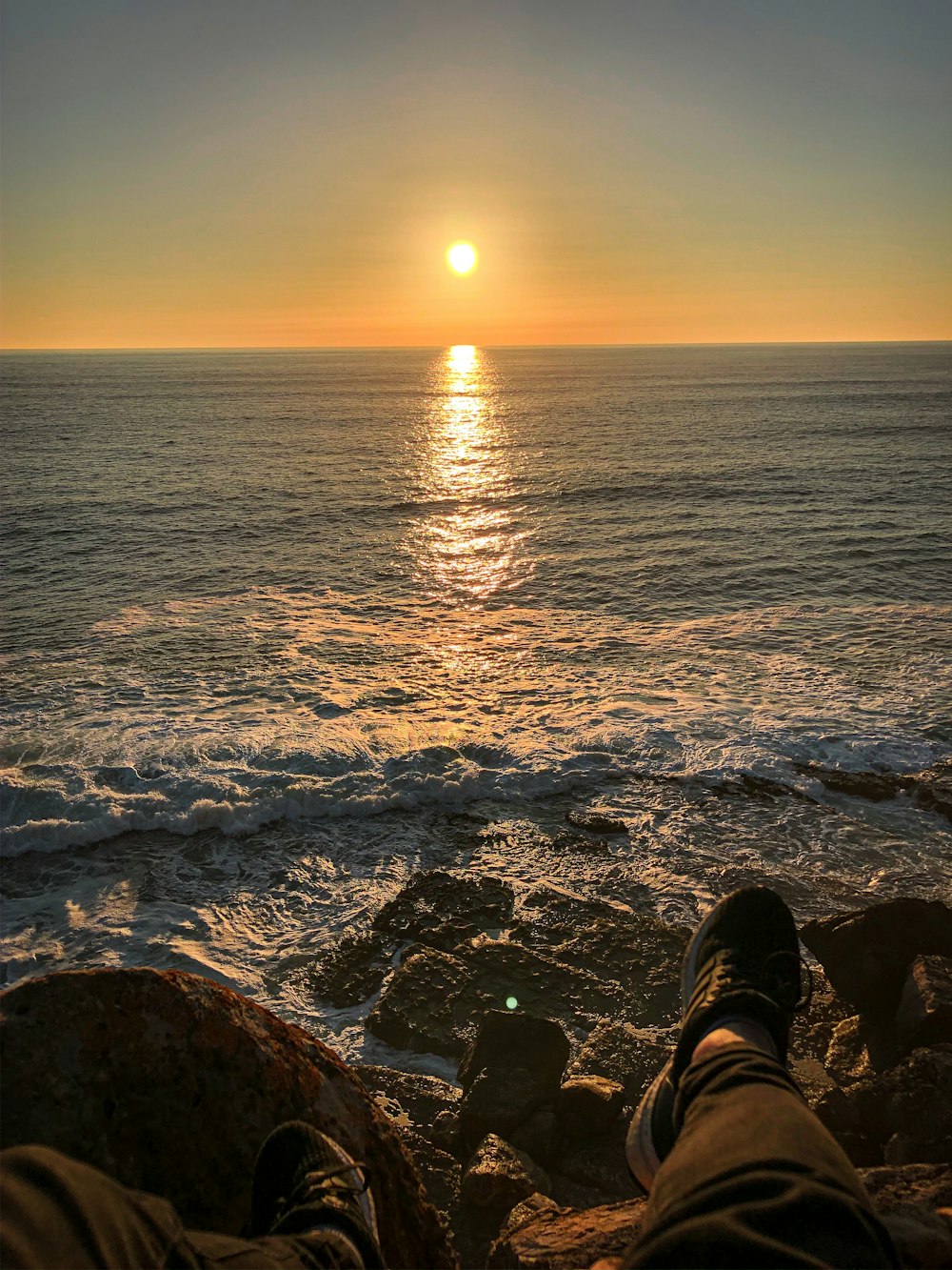 person in black and white sneakers sitting on rock near sea during sunset