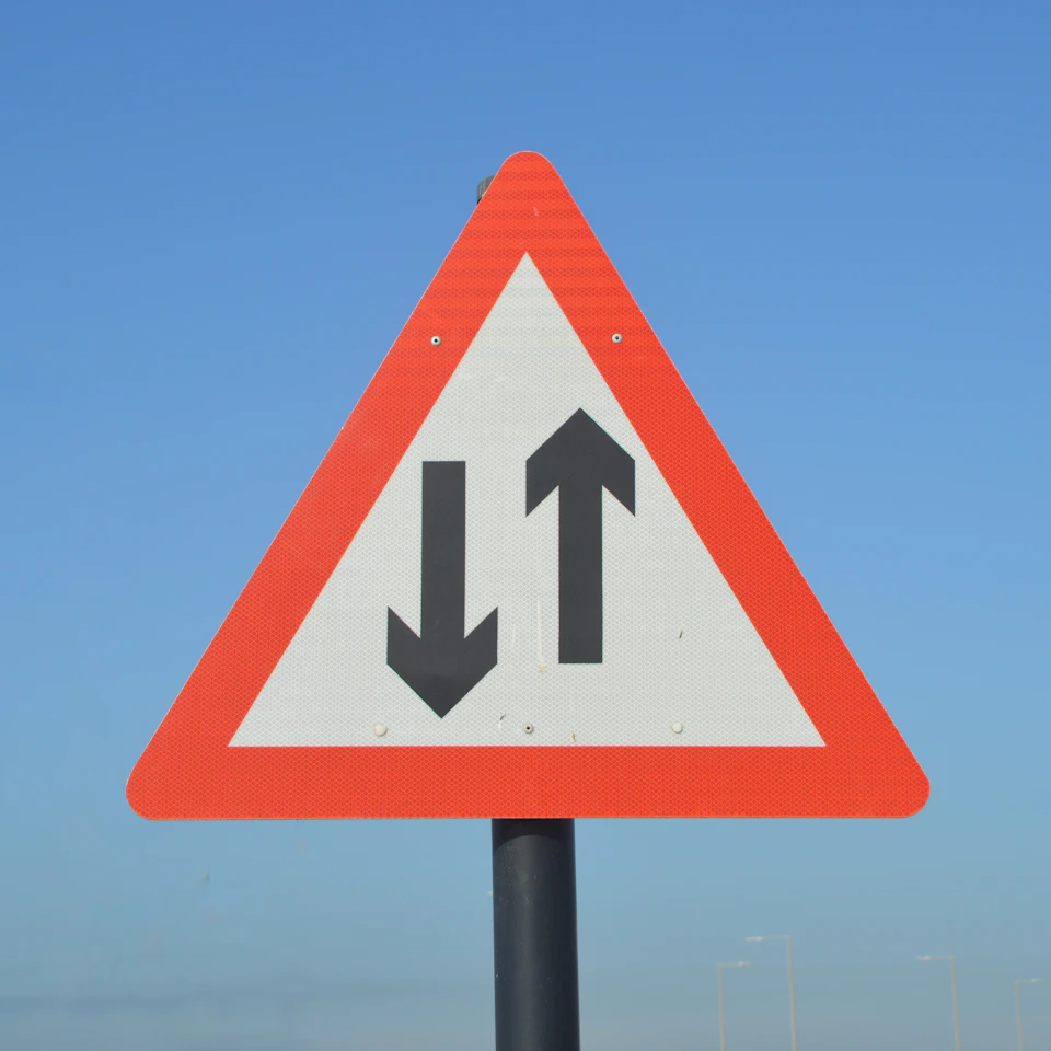 Triangular traffic sign with two black downward-pointing arrows on a roadside pole