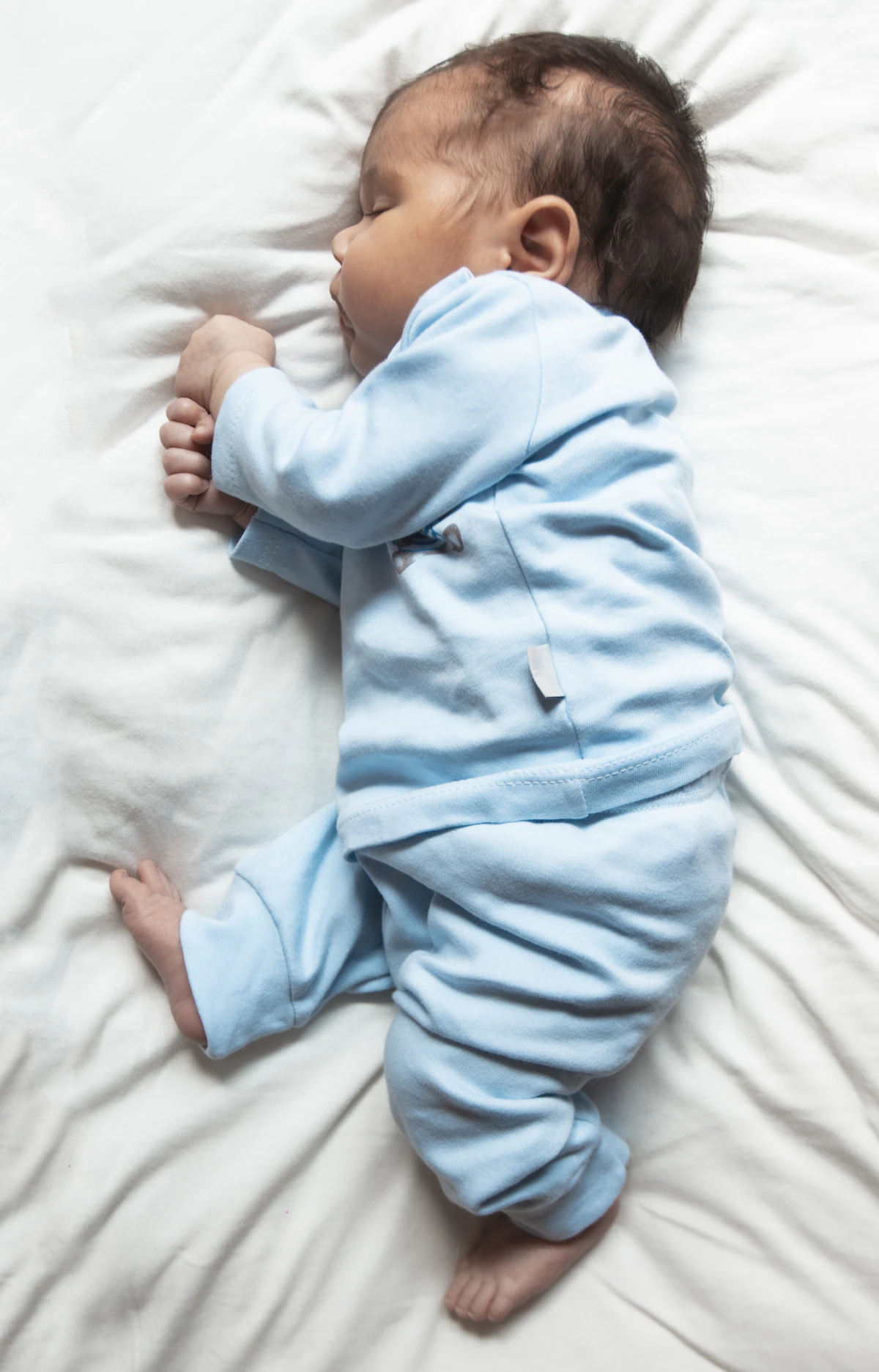 Bebé dormido de lado sobre una sábana blanca con pijama azul