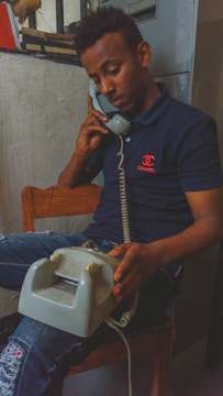 A person is sitting on a wooden chair, holding a vintage rotary telephone to their ear. They are wearing a navy blue polo shirt with a logo and distressed jeans. The background includes a rough, painted wall and part of a window with bars.