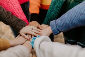 A group of people stand in a circle with their hands stacked on top of each other in the center, suggesting unity and teamwork. They are wearing colorful sweaters.